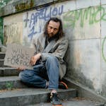 A homeless man sits on steps outdoors holding a 'Will Work for Food' sign, depicting poverty.
