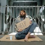 A homeless man sits under a bridge with a sign reading 'HELP', highlighting poverty and homelessness.