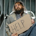 A homeless man with a beard holding a sign asking for help indoors.