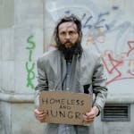 Homeless man standing on the street holding a sign, with graffiti in the background.