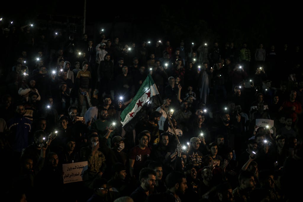 Civil Society Voices people gather at night in Syria, holding flashlights and the Syrian flag, symbolizing unity.