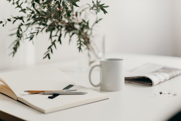 A Lion Newspaper serene minimalist workspace featuring a white coffee mug, open notebook, and green plant vase on a desk.
