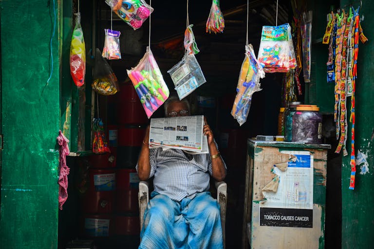 China Daily Web News An elderly man reading a newspaper in a vibrant market shop filled with hanging goods.
