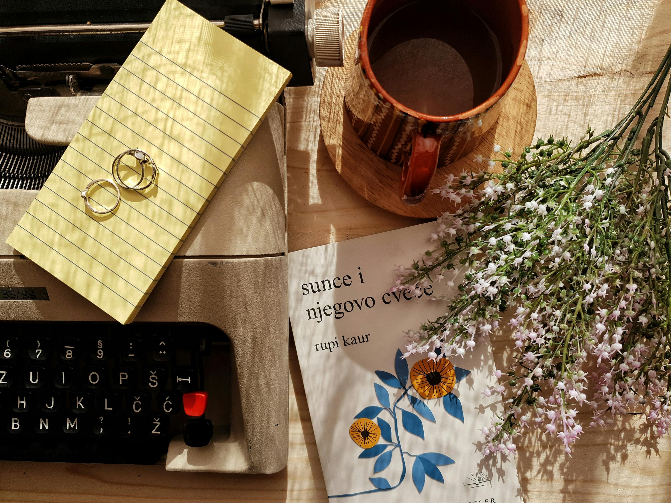 Ravleen Kaur flatlay with a typewriter, coffee mug, flowers, and rings on a wooden surface.