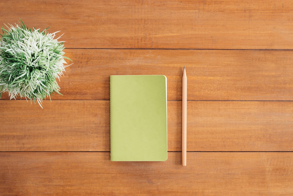 Nerwey Flat lay of green notebook and pencil on wooden desk with plant. Perfect for creativity and organization themes.