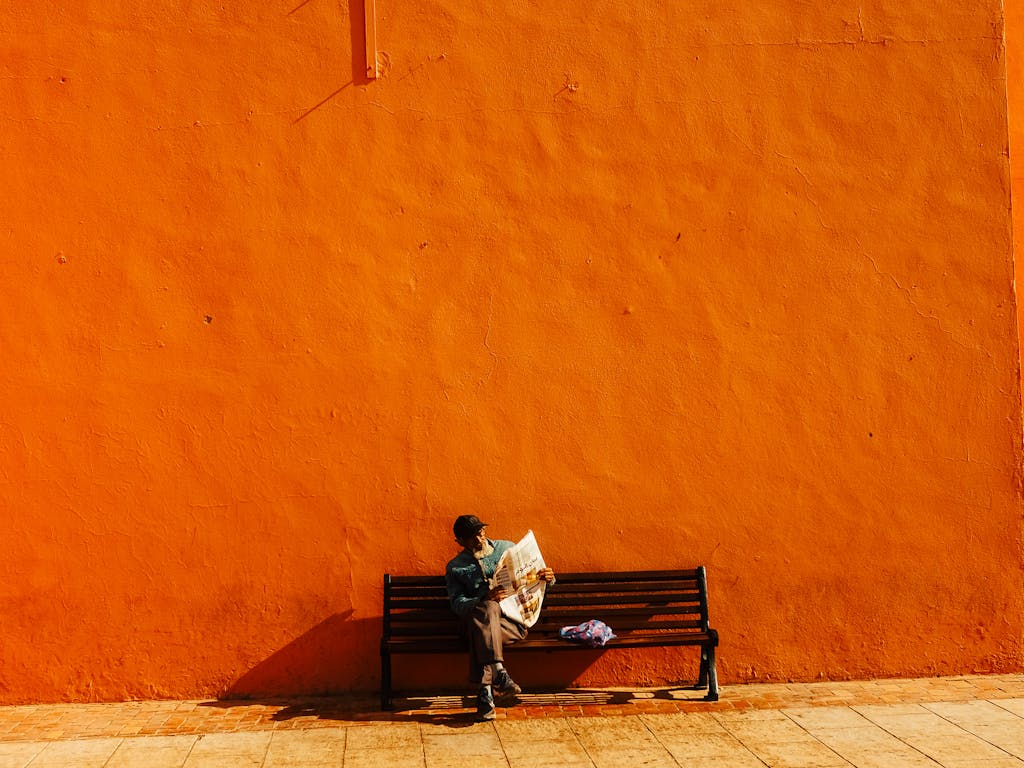 CGTN News Person reading a newspaper on a bench with an orange wall backdrop creates a vivid, minimalist scene.