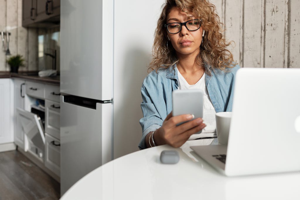 SosoActive News Woman using smartphone and laptop while working remotely in a home kitchen setting.