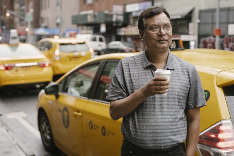 National Breaking News A man enjoys a coffee break beside a yellow taxi in a bustling city street.
