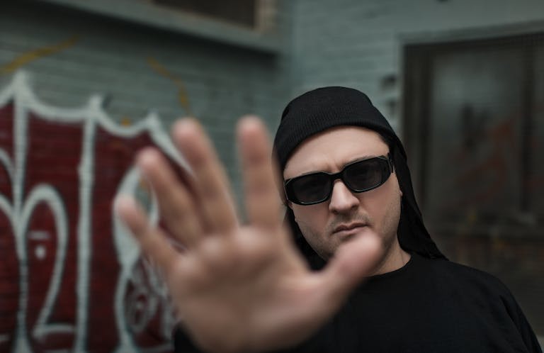 Witeloversb A man in sunglasses puts his hand up in front of the camera against an urban graffiti backdrop.