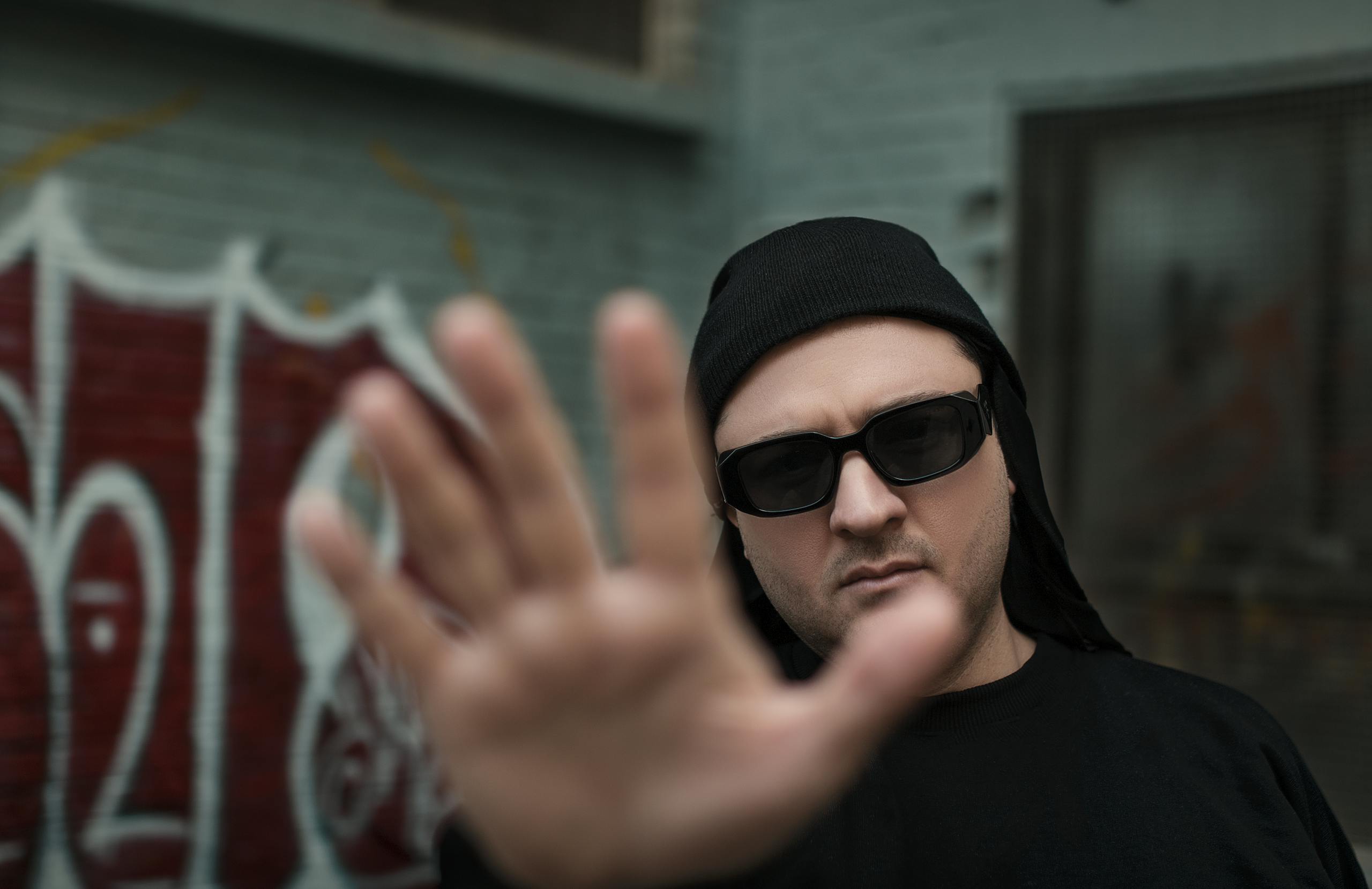 Witeloversb A man in sunglasses puts his hand up in front of the camera against an urban graffiti backdrop.