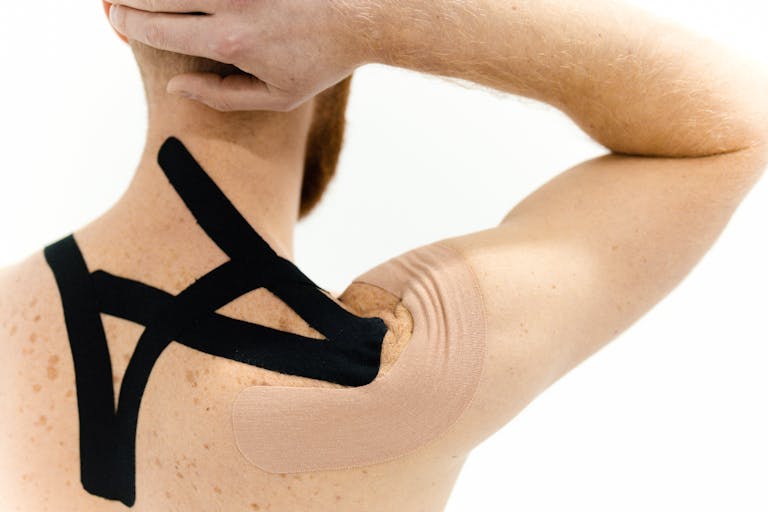 Inomyalgia: Understanding Muscle Pain, Causes, Symptoms, and Effective Management A man with kinesiology tape applied on his shoulder against a white background, showcasing physical therapy support.