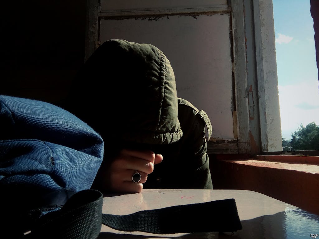 Depomin82 A solitary individual with a hood, seated at a table by an open window, embodying introspection.