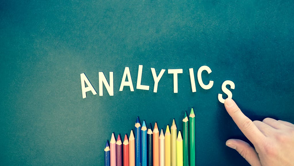 DainikShiksha.com Colored pencils and letters spelling 'ANALYTICS' on a textured background with a hand.