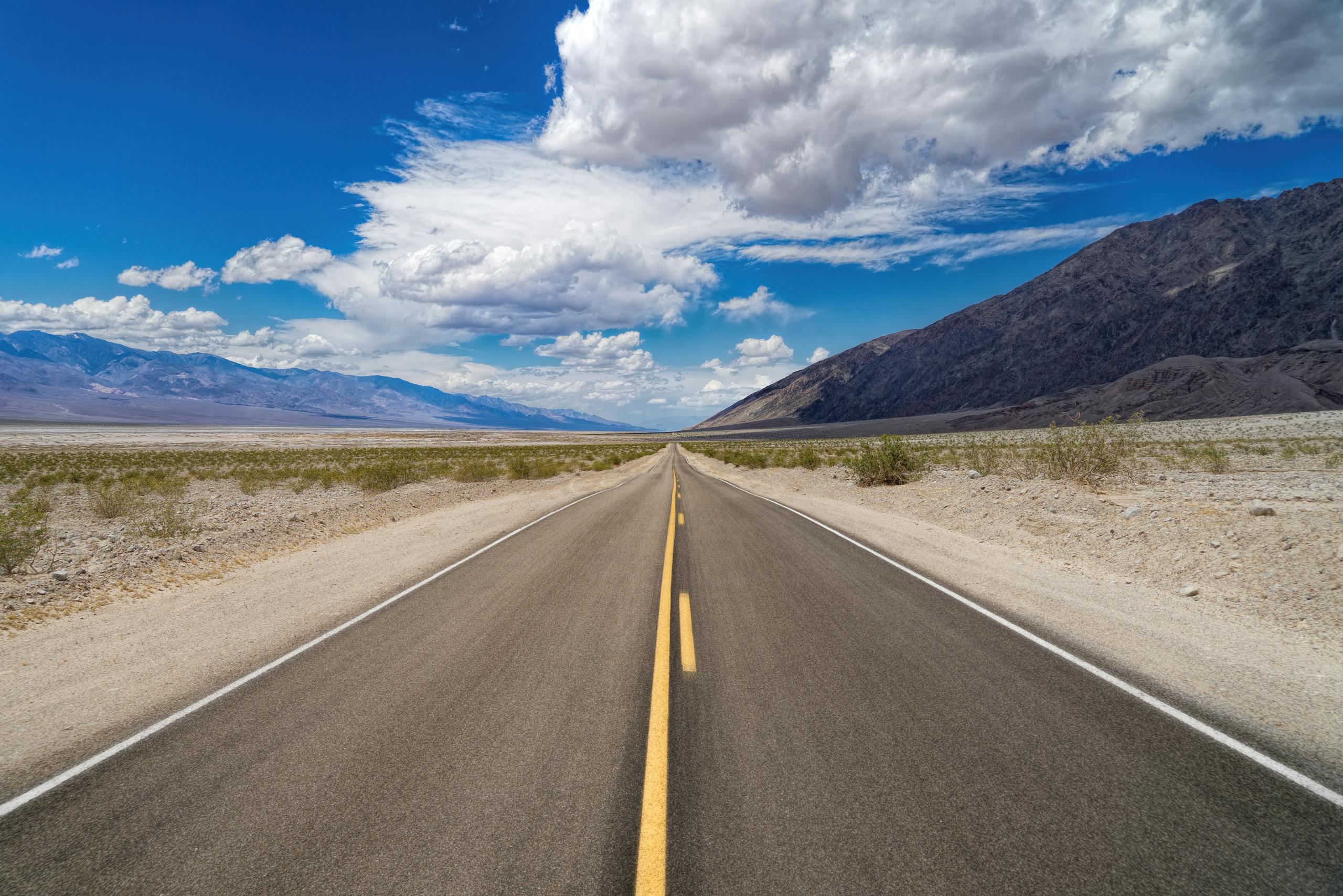 Drivers Death USA Desolate straight road stretching into a desert landscape under a bright blue sky.