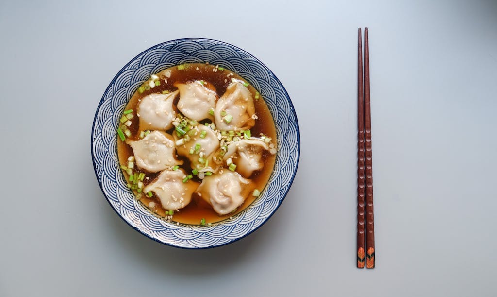 Monika Leveski Top view of a bowl of dumplings in soup garnished with scallions.