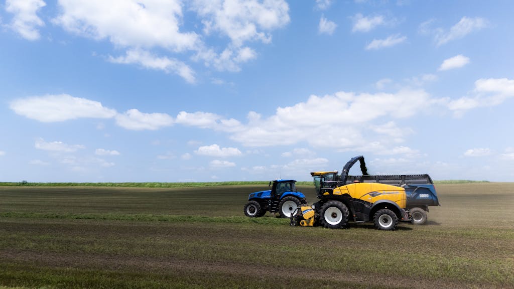 Bin Matcha Two tractors working together in a vast field under a clear blue sky, harvesting crops efficiently.