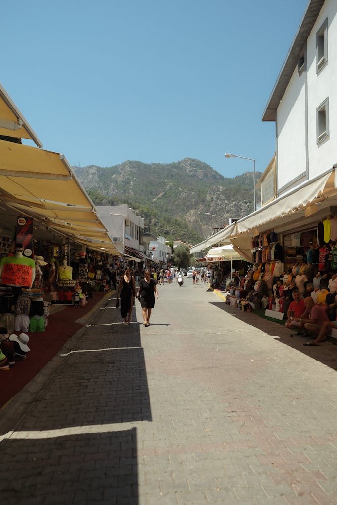 R6 Market Place Lively outdoor market scene with tourists and shoppers, framed by mountains under a clear blue sky.