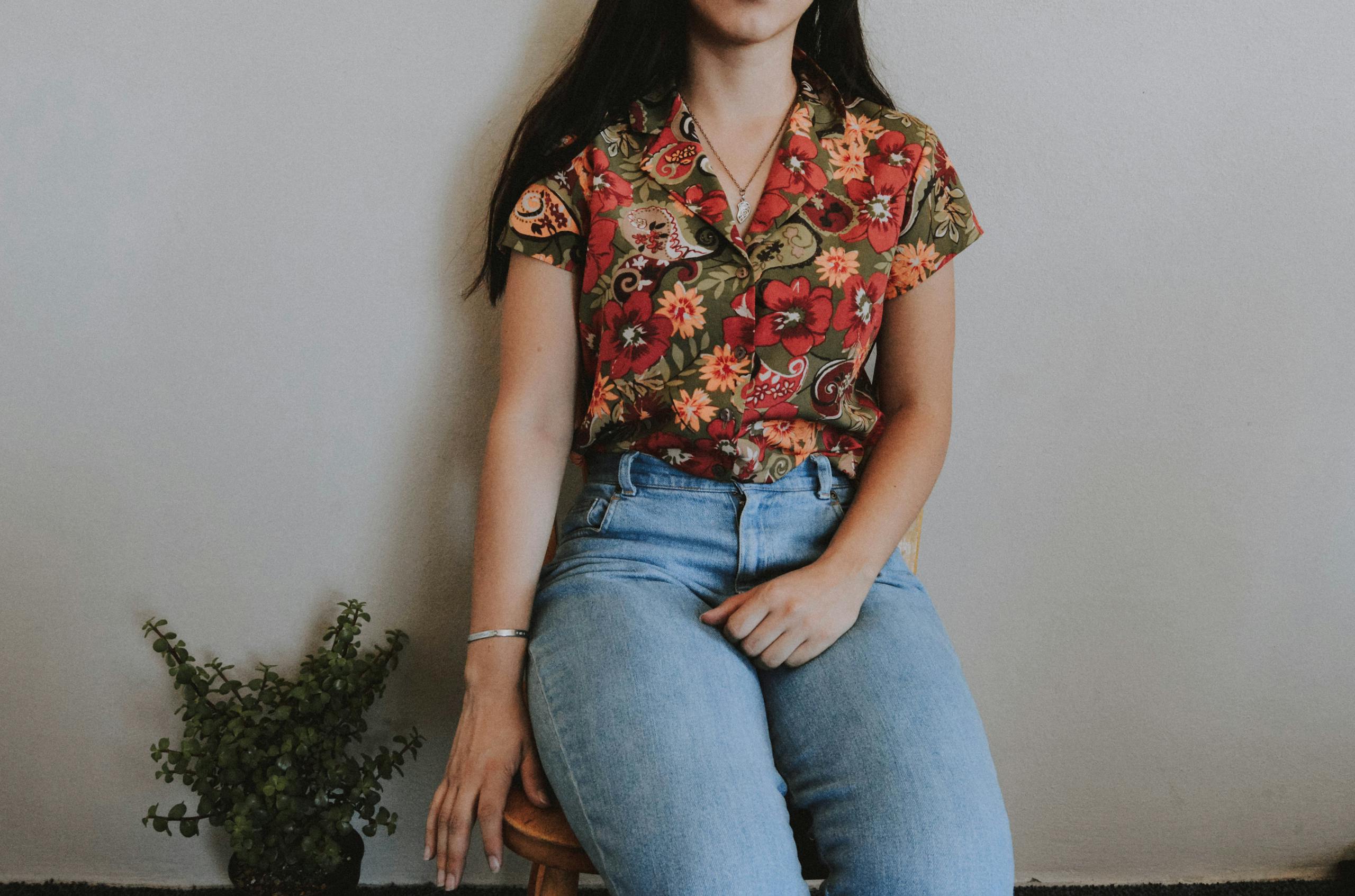 HerbCiepScam Woman sitting on stool wearing floral shirt and jeans indoors with a plant by her side.