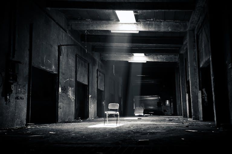 Calesshop A haunting black and white photo of an abandoned warehouse with a solitary chair illuminated by sunlight through a window.