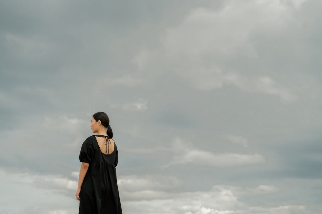 Carmela Clouth  A woman in a black dress stands alone outdoors, gazing at a dramatic cloudy sky.